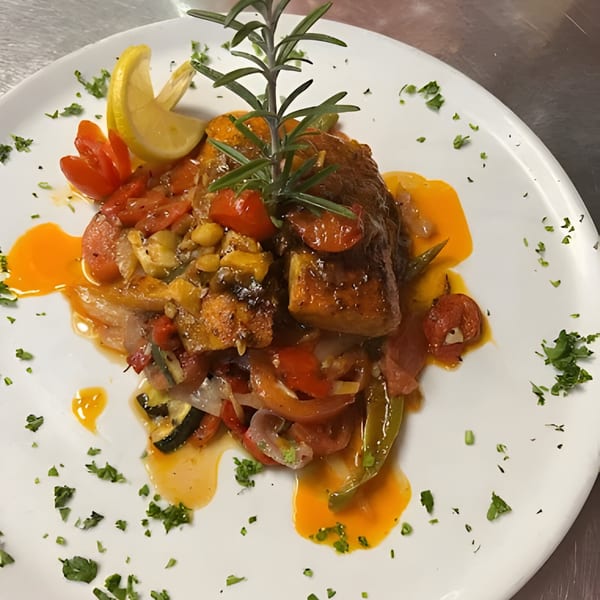 A gourmet Italian dish with fish, roasted vegetables, lemon, and rosemary on a white plate.