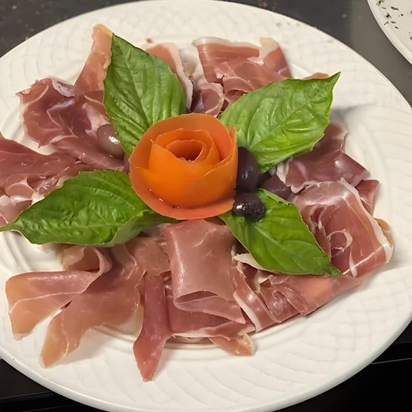 A white plate of thinly sliced prosciutto, garnished with fresh basil leaves and a carved tomato rose.